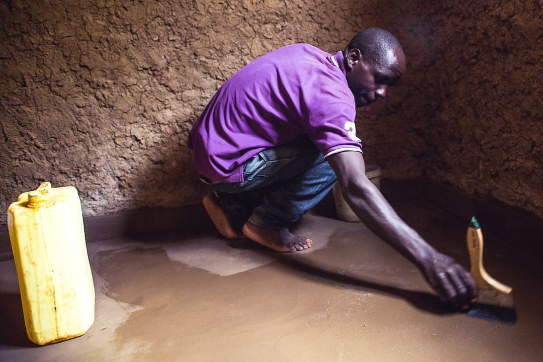 Rwandan man crouched on the ground in a mud hut to install a washable Earth Enable floor.