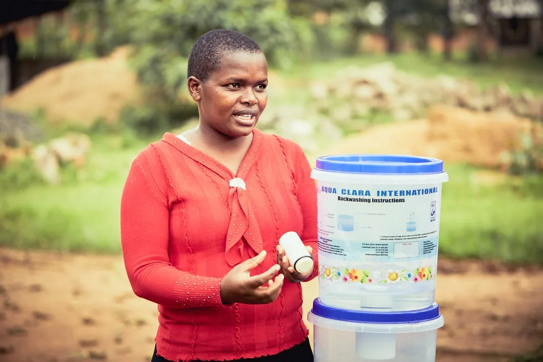 Kenyan woman standing in front of an Aqua Clara water filter setup, explaining to a not shown crowd.