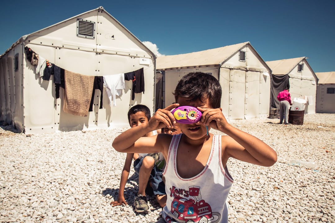 Male Syrian refugee child standing in front of a tent aiming a toy camera at the photographer.