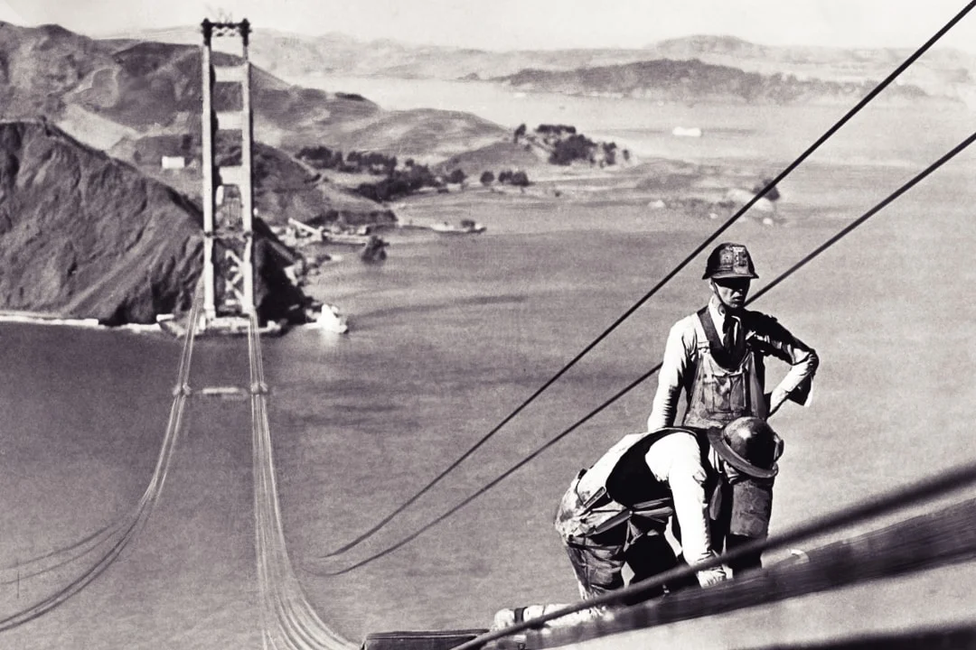 Vintage black and white photo of a man on top of the Golden Gate Bridge during its original construction.