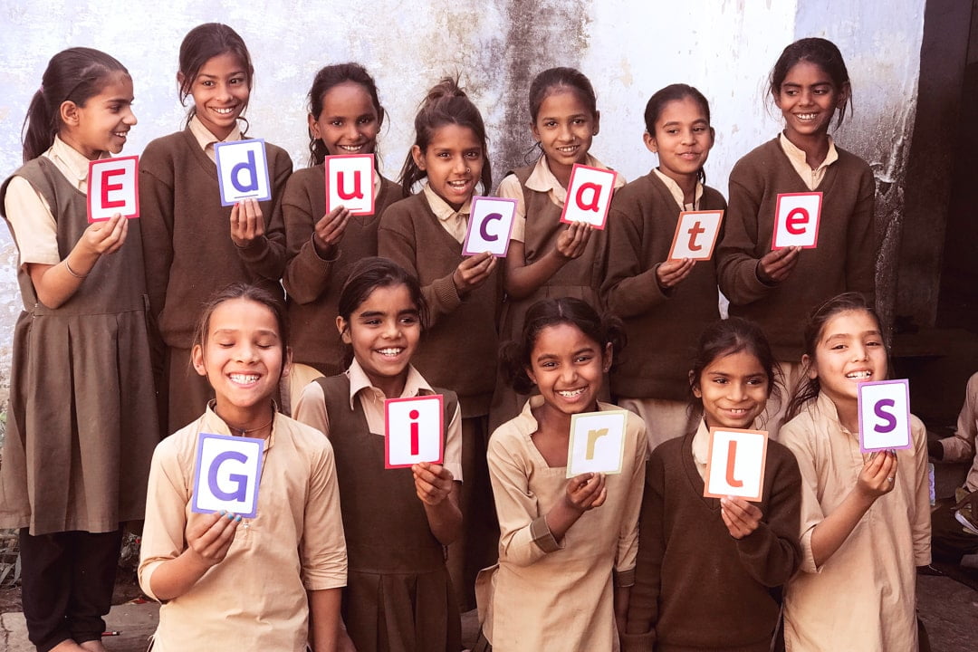 Two rows of young Indian girls in school uniforms, each holding a sign with a different letter to spell out Educate Girls.