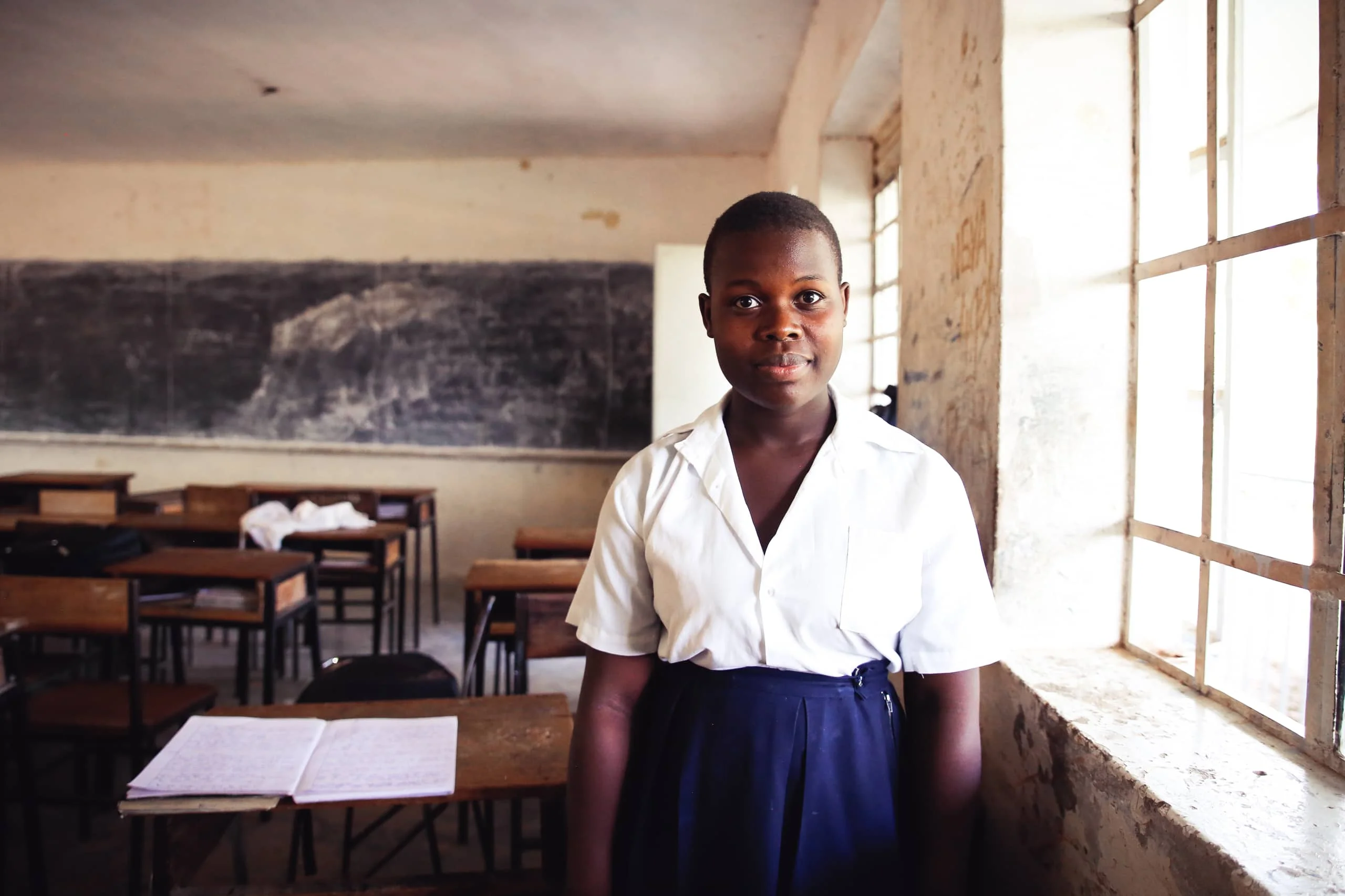 Female Ugandan secondary student in school uniform standing by a window in front of an empty classroom and blackboard