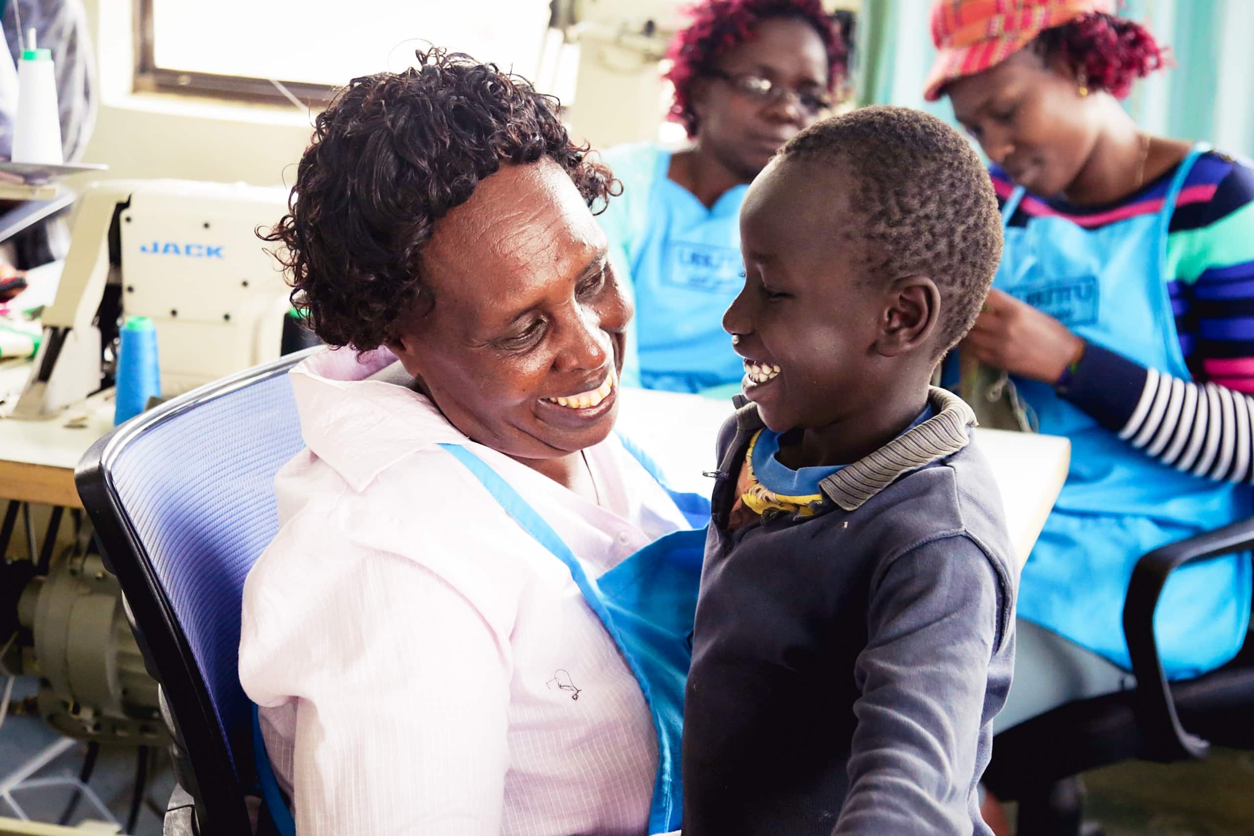 Kenyan mother holding and smiling at her young male son in the factory where she creates shoes for Ubuntu Life.