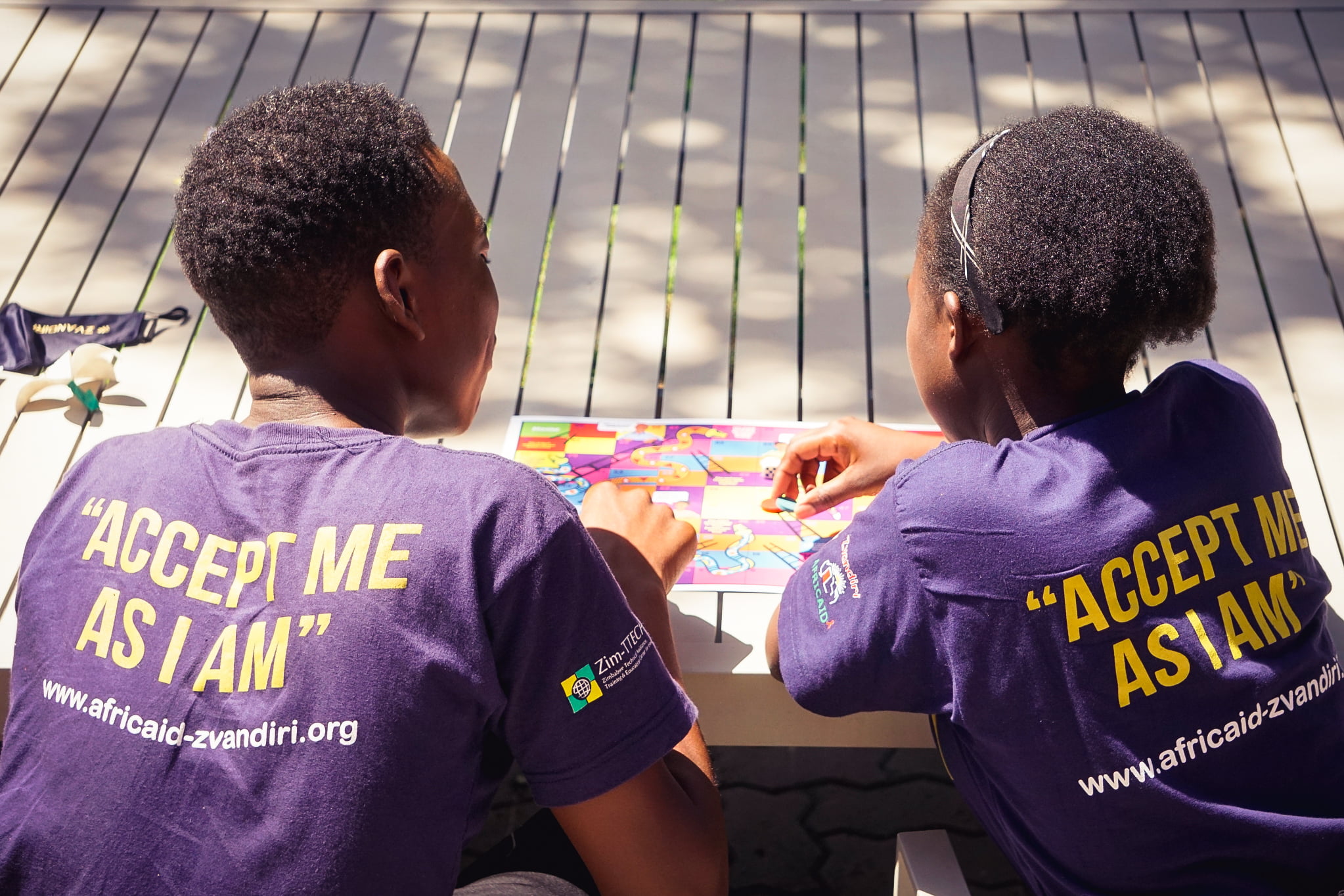 A male and female African youth playing a board game, wearing t-shirts that read "Accept me as I am."