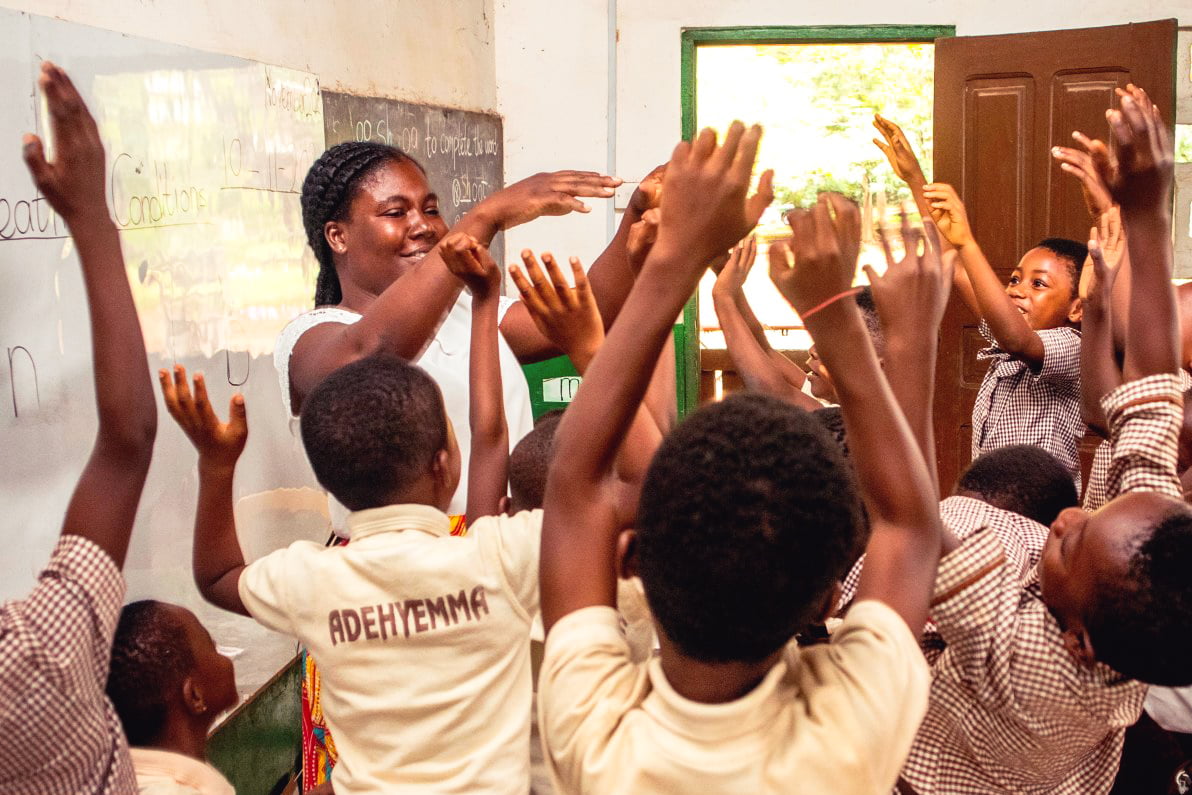 African female teacher teaching a group of enthusiastic learners with their hands up.